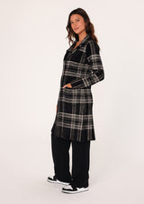 [Color: Black/Cream] A side facing image of a brunette model wearing a cozy and fuzzy sweater coat in a black and cream plaid pattern. With long sleeves, a notched lapel, side patch pockets, and a button front. 