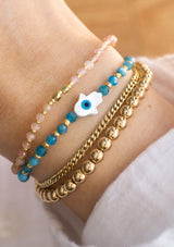 [Title: Apatite]  An adjustable healing bracelet, crafted on a durable nylon cord with a mother of pearl hamsa stone, gold-plated hardware and teal blue natural gemstones. Designed by Kindred Row.