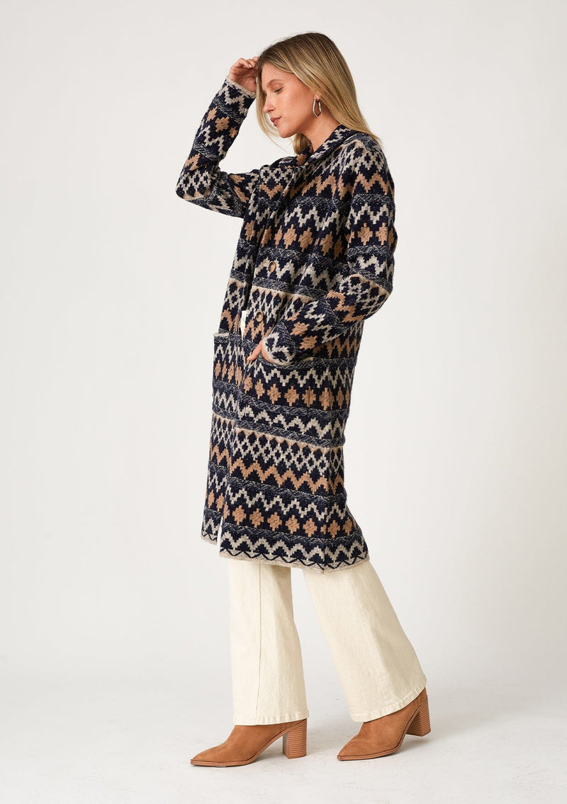 [Color: Navy/Camel] A side facing image of a blonde model wearing a sweater coat in a bohemian blue and brown chevron striped design. With long sleeves, a notched collar, a button front, side patch pockets, and a back vent. 
