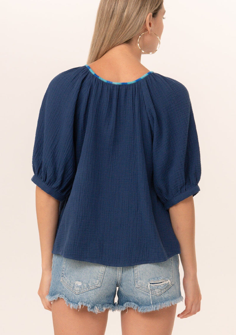 [Color: Navy] A back facing image of a blonde model wearing a navy blue cotton gauze blouse. With short raglan puff sleeves, a round neckline with contrast thread details, and a button front.