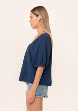 [Color: Navy] A side facing image of a blonde model wearing a navy blue cotton gauze blouse. With short raglan puff sleeves, a round neckline with contrast thread details, and a button front.