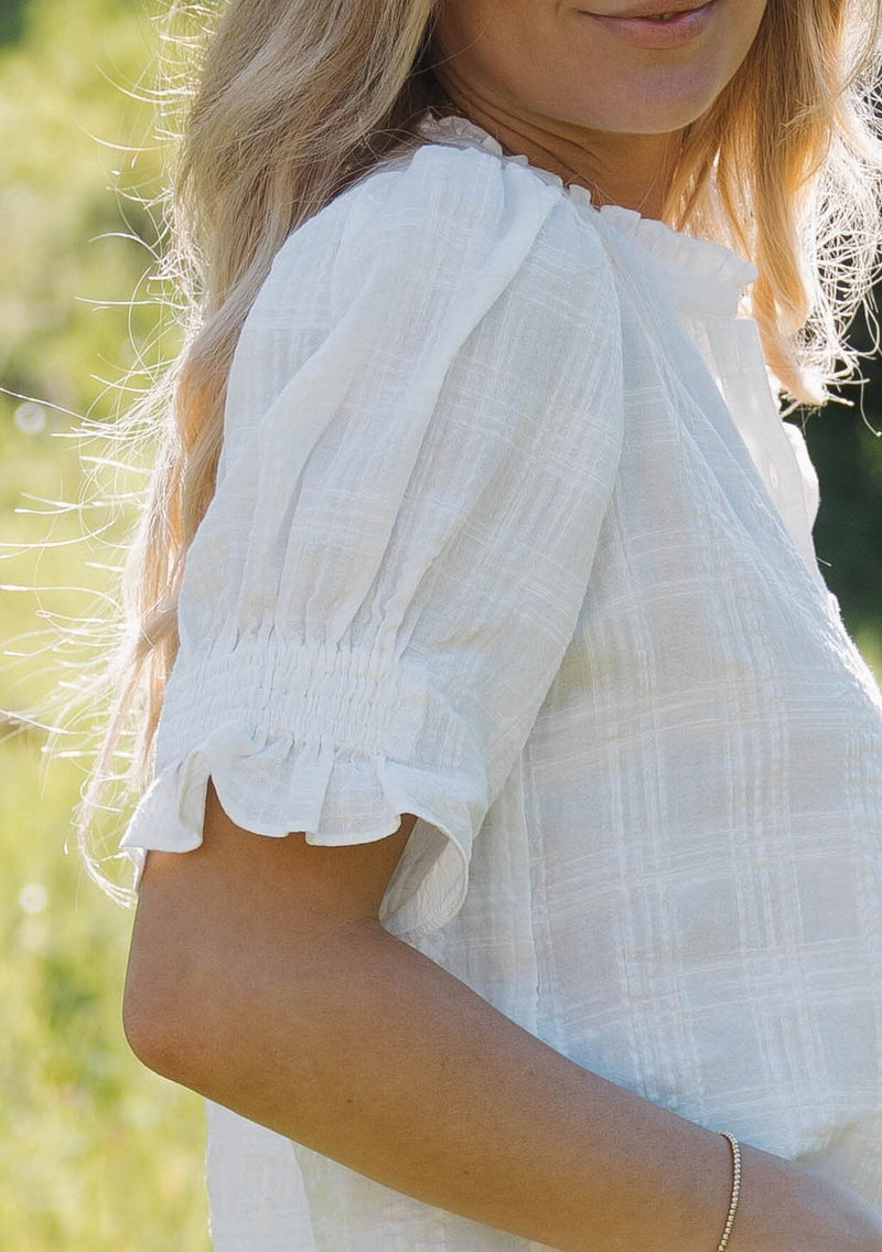 [Color: White] A front facing image of a blonde model wearing a white bohemian cotton blend blouse in a textured gingham. With short puff sleeves, a button front, a high ruffled neckline, and smocked elastic details at the sleeve.