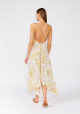 [Color: Dusty Peach/Caramel] A back facing image of a brunette model wearing a peach pink bohemian halter dress with paisley print details and gold metallic clip dot details. With adjustable spaghetti straps that can be tied in multiple ways, a sexy open back, a v neckline with mini pom trim, and an asymmetric handkerchief hemline. 