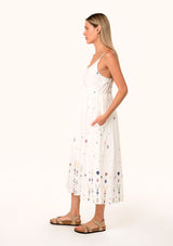 [Color: Natural/Olive] A front facing image of a blonde model wearing a sleeveless white cotton bohemian mid length dress. With adjustable spaghetti straps, a scooped neckline, side pockets, and multi colored embroidered details throughout. 