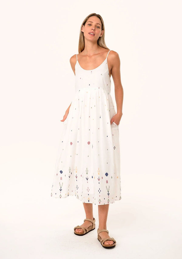 [Color: Natural/Olive] A front facing image of a blonde model wearing a sleeveless white cotton bohemian mid length dress. With adjustable spaghetti straps, a scooped neckline, side pockets, and multi colored embroidered details throughout. 