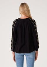 [Color: Black/Natural] A back facing image of a blonde model wearing a bohemian black blouse with embroidered detail. With voluminous long sleeves, a v neckline, and a relaxed fit.