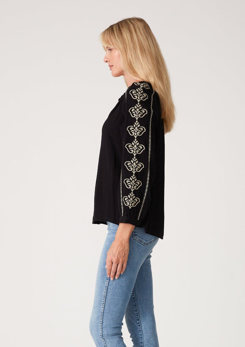 [Color: Black/Natural] A side facing image of a blonde model wearing a bohemian black blouse with embroidered detail. With voluminous long sleeves, a v neckline, and a relaxed fit.