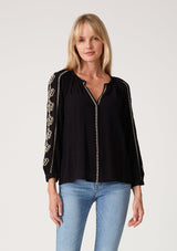 [Color: Black/Natural] A front facing image of a blonde model wearing a bohemian black blouse with embroidered detail. With voluminous long sleeves, a v neckline, and a relaxed fit.