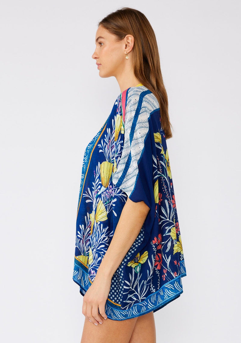 [Color: Navy/Yellow] A side facing image of a brunette model wearing a bohemian style kimono top in a navy blue and yellow floral print with a contrast border. A lightweight beach cover up style with half length sleeves, an open front, and a hip length hemline. 