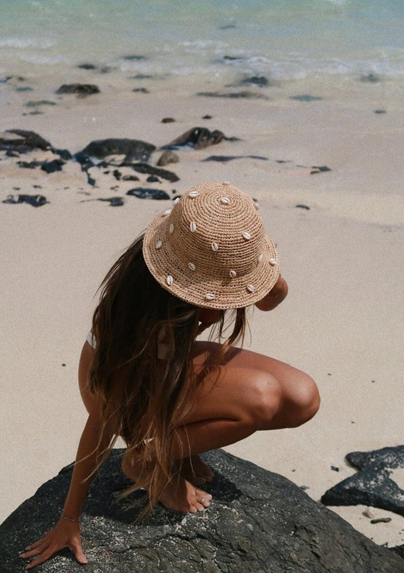 A blonde model wearing a raffia bucket hat with sewn in cowry shells.