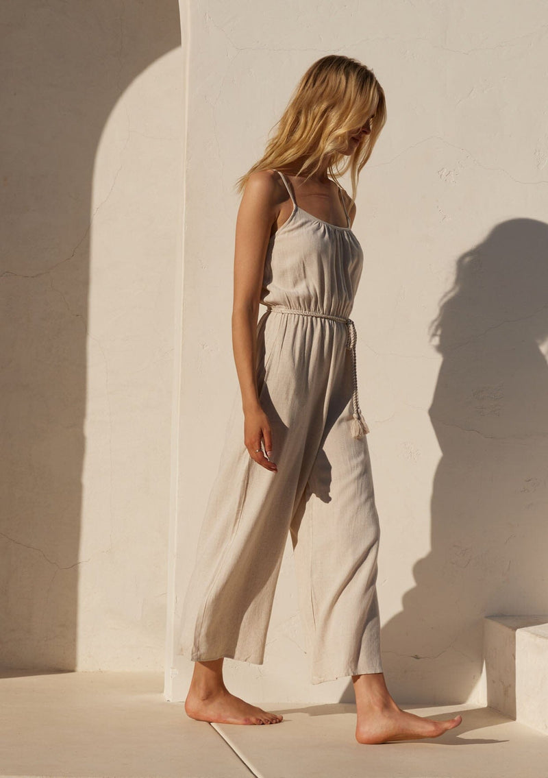 [Color: Natural] A side facing image of a blonde model wearing an off white sleeveless jumpsuit. With adjustable spaghetti straps, a scooped neckline, a long wide leg, side pockets, an elastic waist, and a braded tassel belt.