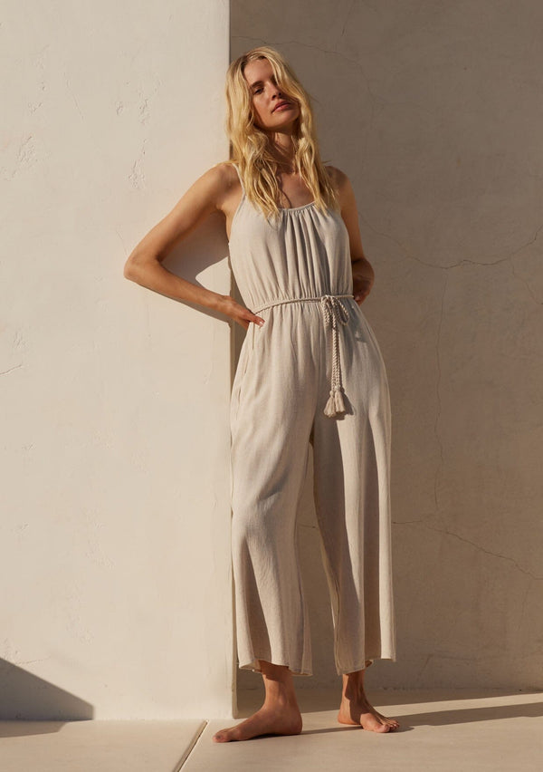 [Color: Natural] A full body front facing image of a blonde model wearing an off white sleeveless jumpsuit. With adjustable spaghetti straps, a scooped neckline, a long wide leg, side pockets, an elastic waist, and a braded tassel belt.