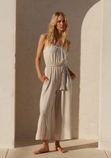 [Color: Natural] A front facing image of a blonde model wearing an off white sleeveless jumpsuit. With adjustable spaghetti straps, a scooped neckline, a long wide leg, side pockets, an elastic waist, and a braded tassel belt.
