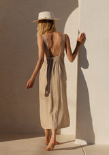 [Color: Wheat] A back facing image of a blonde model wearing a loose fit mid length dress in a light khaki linen blend. With thick tank top straps, a low back, and side pockets.