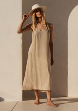 [Color: Wheat] A front facing image of a blonde model wearing a loose fit mid length dress in a light khaki linen blend. With thick tank top straps, a low back, and side pockets.