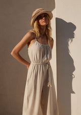 [Color: Natural] A half body front facing image of a blonde model wearing an off white sleeveless jumpsuit. With adjustable spaghetti straps, a scooped neckline, a long wide leg, side pockets, an elastic waist, and a braded tassel belt.