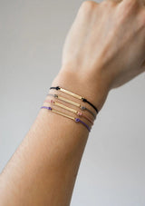 [Color: Onyx] A close-up of a person's wrist wearing four delicate charm bracelets stacked together. The bracelets include gold, rose gold, pink, and black cords with small decorative charms, set against a plain light gray background.