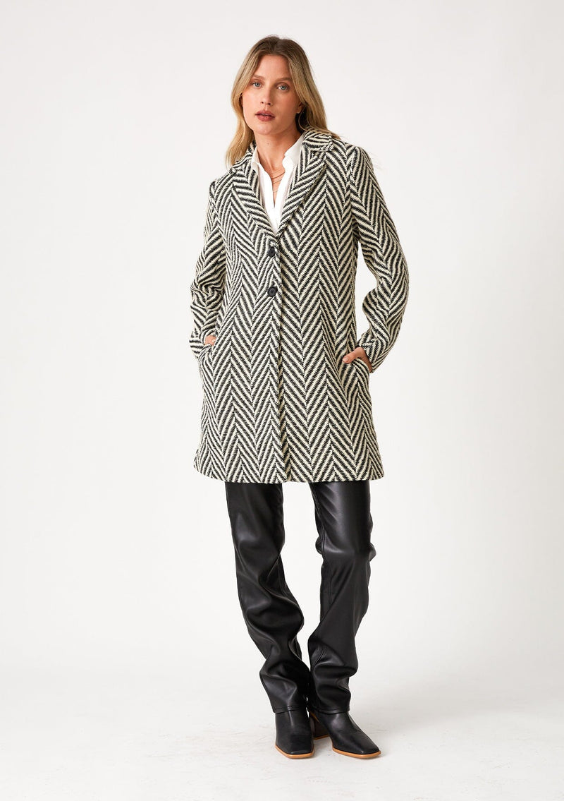 [Color: Cream/Black] A full body front facing image of a blonde model wearing a classic womens coat in a cream and black chevron stripe. With long sleeves, a notched collar, a button front, and side pockets. 