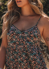 [Color: Black/Teal] A front facing image of a blonde model standing outside wearing a bohemian maxi dress in a black and teal floral print. With adjustable spaghetti straps, a scalloped trim v neckline with contrast thread details, a tiered flowy silhouette, side pockets, and an empire waist.