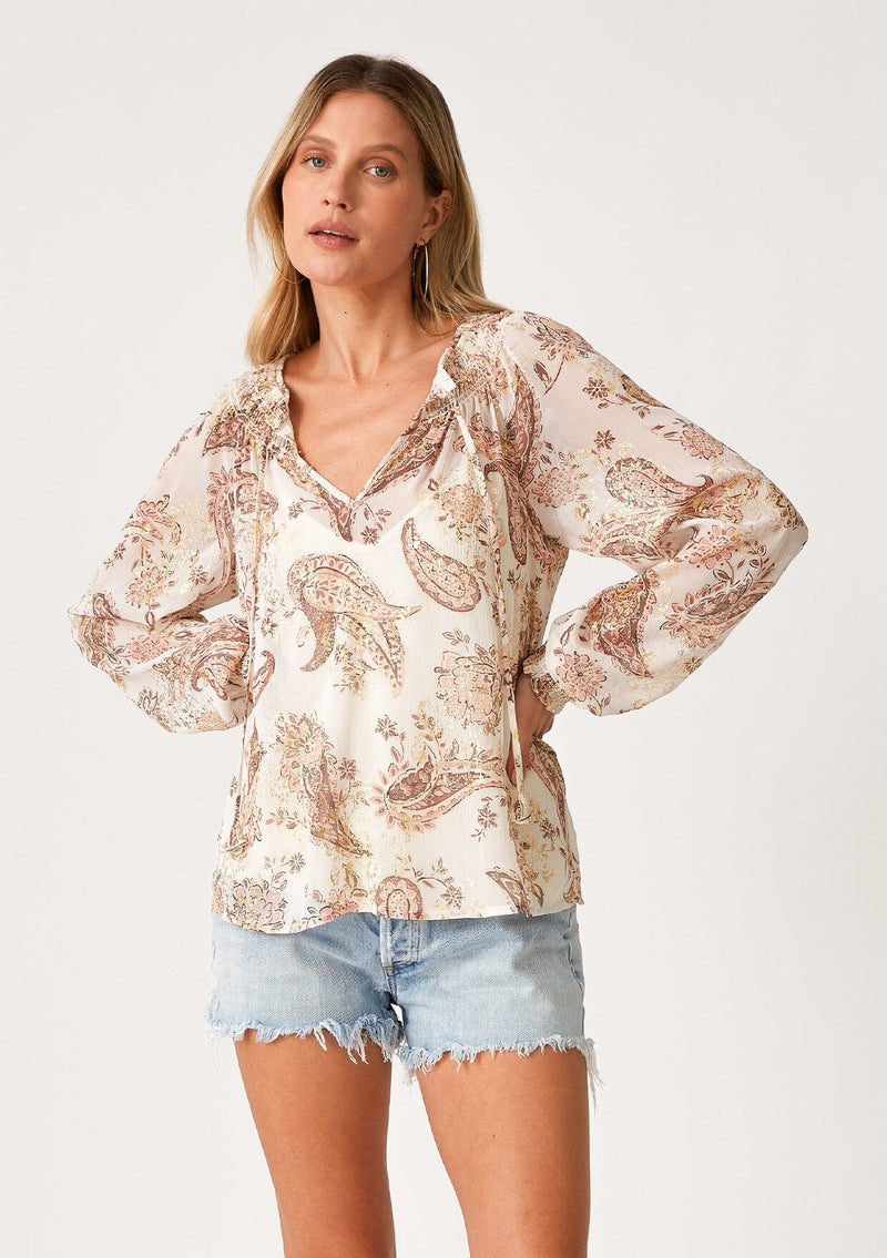 [Color: Natural/Dusty Rose] A half body front facing image of a blonde model wearing a fall bohemian blouse in an ivory and dusty pink paisley print. With gold metallic details throughout, three quarter length raglan sleeves, ruffled elastic wrist cuffs, a smocked neck, and a v neckline with ties. 