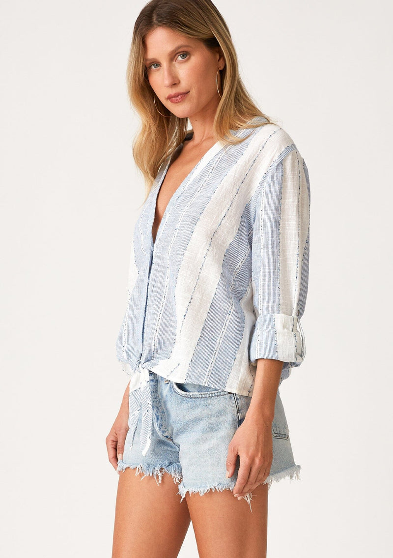 [Color: White/Blue] A side facing image of a blonde model wearing a lightweight white and blue striped top. With long rolled sleeves, a tie front detail, and a concealed button up front.