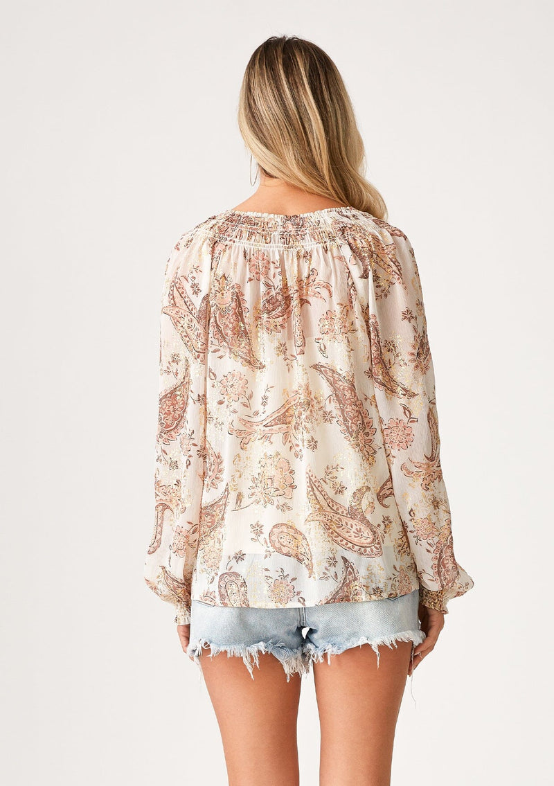 [Color: Natural/Dusty Rose] A back facing image of a blonde model wearing a fall bohemian blouse in an ivory and dusty pink paisley print. With gold metallic details throughout, three quarter length raglan sleeves, ruffled elastic wrist cuffs, a smocked neck, and a v neckline with ties. 