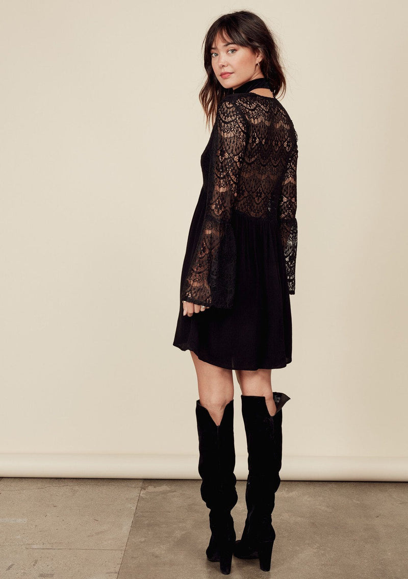 [Color: Black] A brunette woman wearing a sheer lace mini dress. Featuring a feminine v neckline, a button front detail, an empire waist, long sheer lace bell sleeves, and a sexy sheer lace back detail.