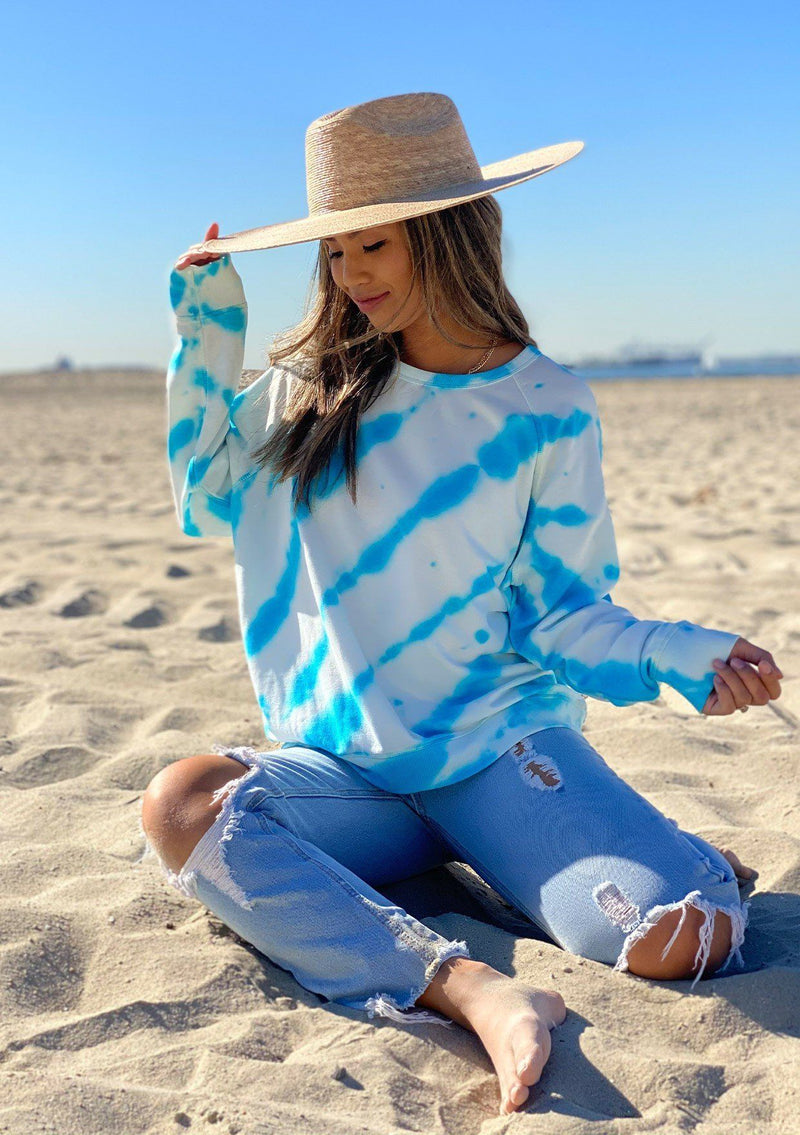 [Color: White Lagoon] A woman on the beach wearing a soft and cozy tie dye sweatshirt. This cozy pullover features long raglan sleeves and a classic crew neckline. A great option for working from home.