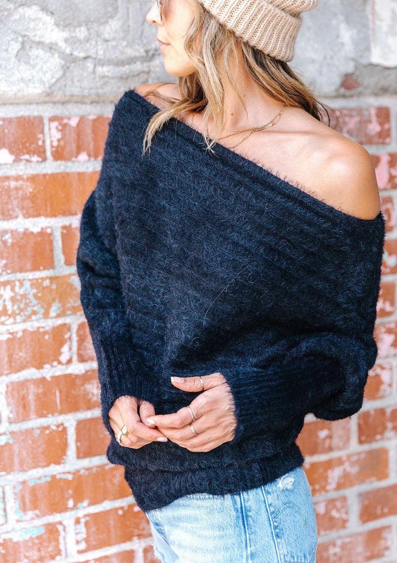 [Color: Black] A blonde model wearing a fuzzy black boatneck sweater. The wide neckline can be worn off the shoulder.  