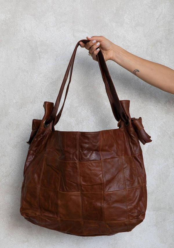 [Color: Brown] A brown leather bag with patchwork stitch exterior, double leather straps, and o ring detail. 