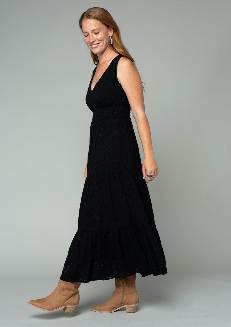[Color: Black] A side facing image of a red headed model wearing a sleeveless black bohemian eyelet maxi dress. With a v neckline, a long tiered skirt, and a smocked elastic waist. A classic bohemian black maxi dress for Summer.