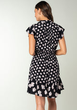 [Color: Black/Taupe] A back facing image of a brunette model wearing a bohemian black mini dress in a taupe floral print. With short ruffled sleeves, a tiered flowy skirt, and a button front. 