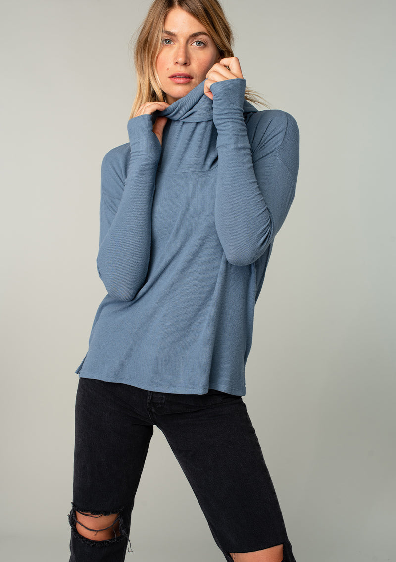 [Color: Denim] A model wearing a soft bamboo micro rib long sleeve top. Featuring an exaggerated cowl neckline that doubles as a hood, long sleeves with thumbhole accents, and breezy side vents.