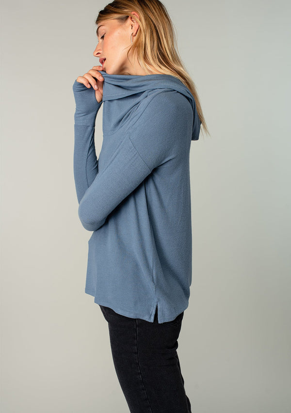 [Color: Denim] A model wearing a soft bamboo micro rib long sleeve top. Featuring an exaggerated cowl neckline that doubles as a hood, long sleeves with thumbhole accents, and breezy side vents.