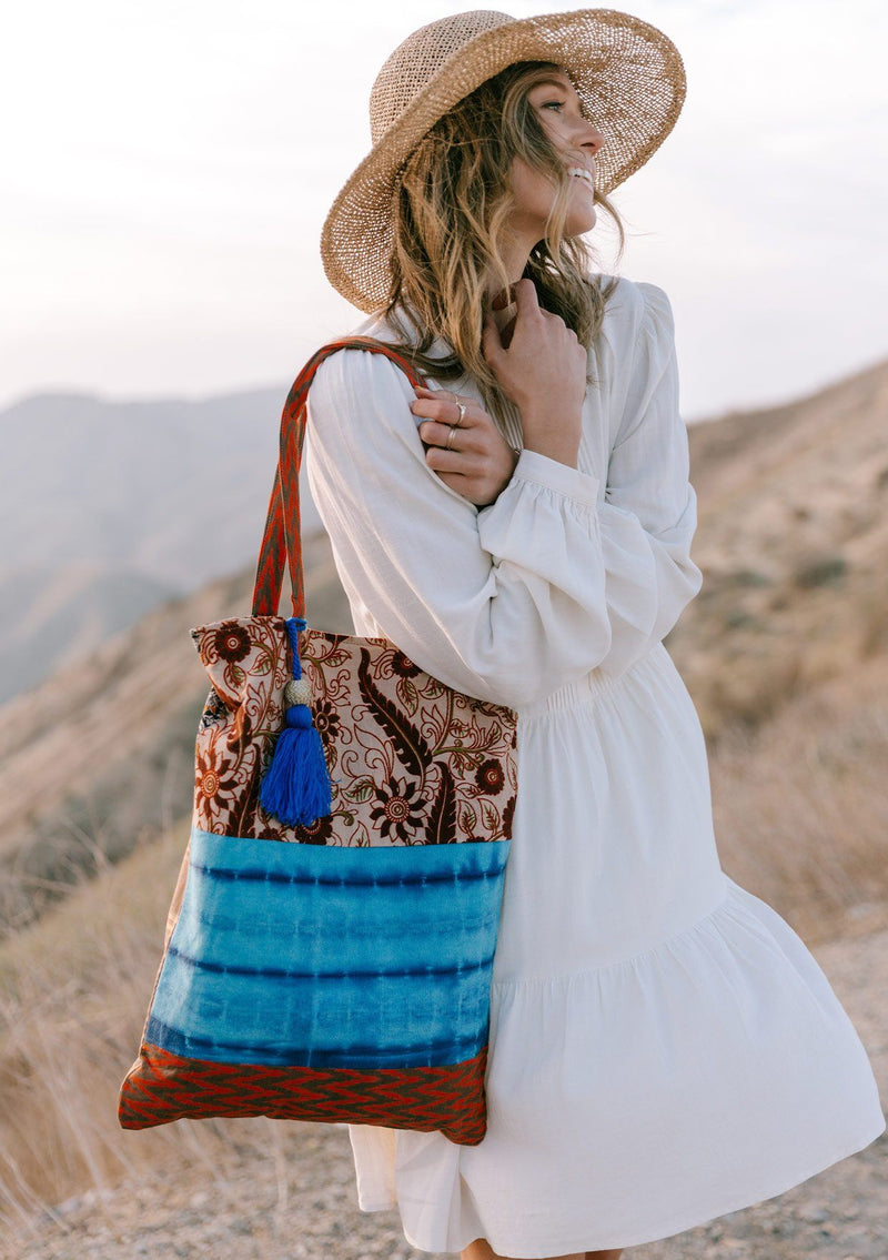 [Color: Natural/Blue] A woman standing outside on a cliff wearing a white dress and holding a bohemian tie dye patchwork tote bag. Featuring a large tassel accent. 