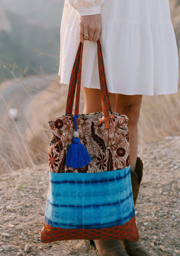 [Color: Natural/Blue] A woman standing outside on a cliff wearing a white dress and holding a bohemian tie dye patchwork tote bag. Featuring a large tassel accent. 