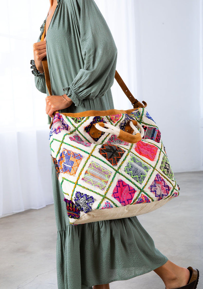 [Color: Multi/Natural] A model holding a patchwork weekender bag. With embroidered details, suede trim, suede top handles, and a suede long adjustable strap.