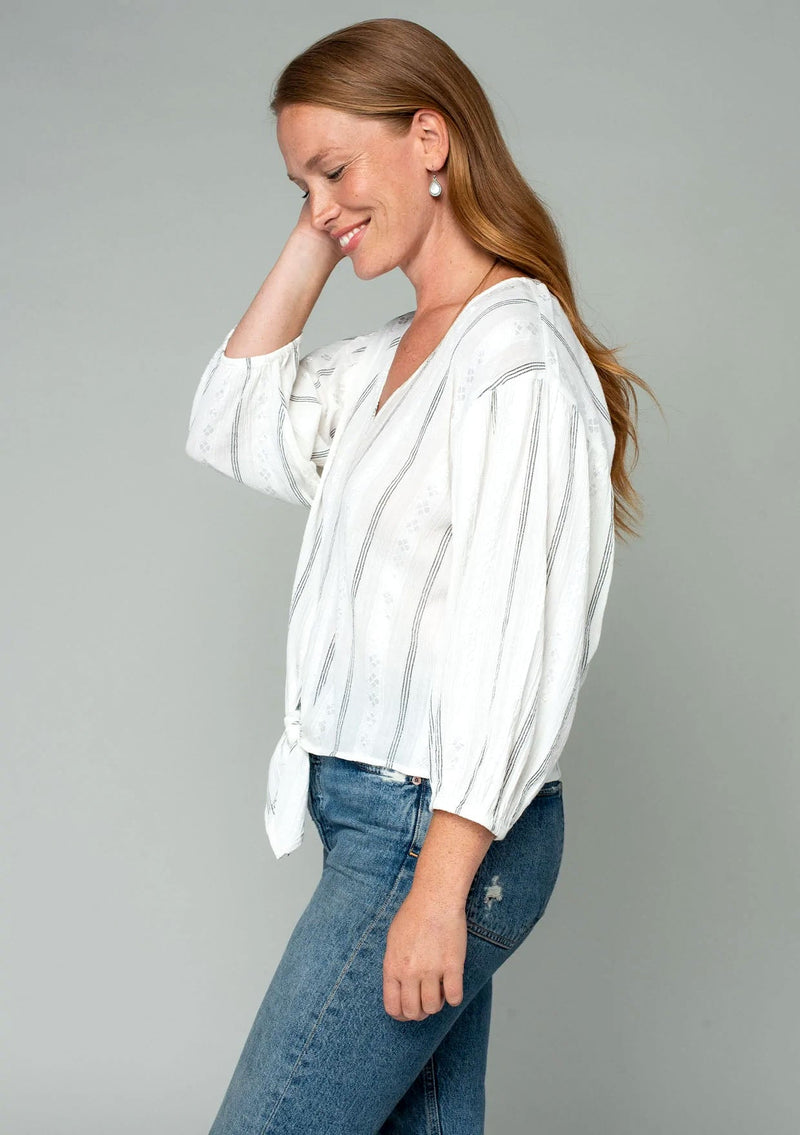 [Color: Off White/Black] A front facing image of a red headed model wearing a white long sleeve bohemian top with a thin black yarn dye stripe throughout. With a v neckline and a tie waist detail. 