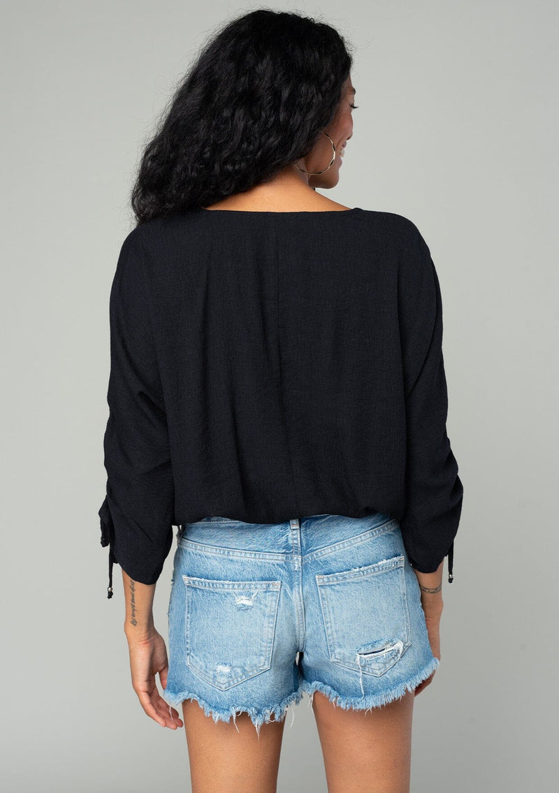 [Color: Black] A back facing image of a brunette model wearing a classic black bohemian top with a self covered button front, a v neckline, and three quarter length sleeves with a gathered sleeve detail and adjustable ties. 