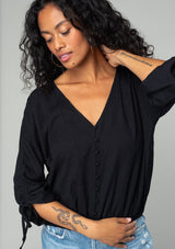 [Color: Black] A close up front facing image of a brunette model wearing a classic black bohemian top with a self covered button front, a v neckline, and three quarter length sleeves with a gathered sleeve detail and adjustable ties. 