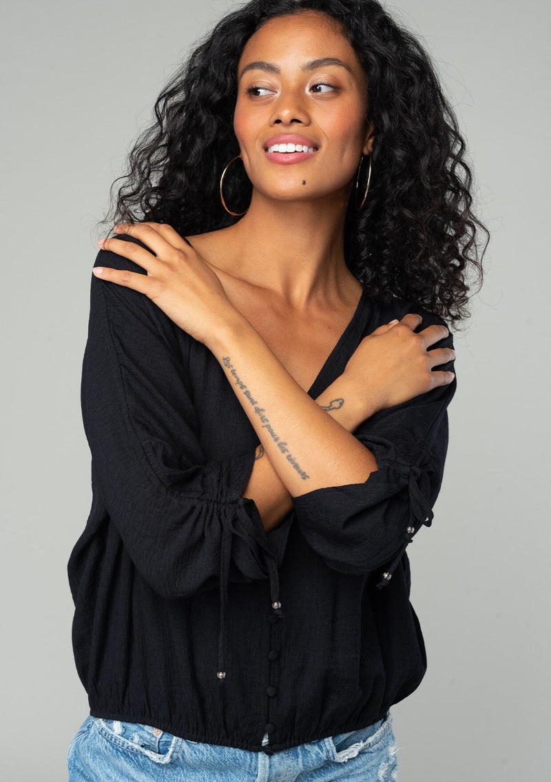 [Color: Black] A front facing image of a brunette model wearing a classic black bohemian top with a self covered button front, a v neckline, and three quarter length sleeves with a gathered sleeve detail and adjustable ties. 