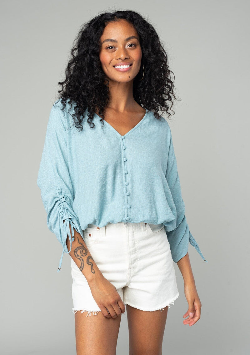 [Color: Dusty Blue] A front facing image of a brunette model wearing a classic dusty blue bohemian top with a self covered button front, a v neckline, and three quarter length sleeves with a gathered sleeve detail and adjustable ties. 