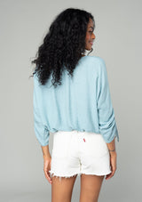[Color: Dusty Blue] A back facing image of a brunette model wearing a classic dusty blue bohemian top with a self covered button front, a v neckline, and three quarter length sleeves with a gathered sleeve detail and adjustable ties. 