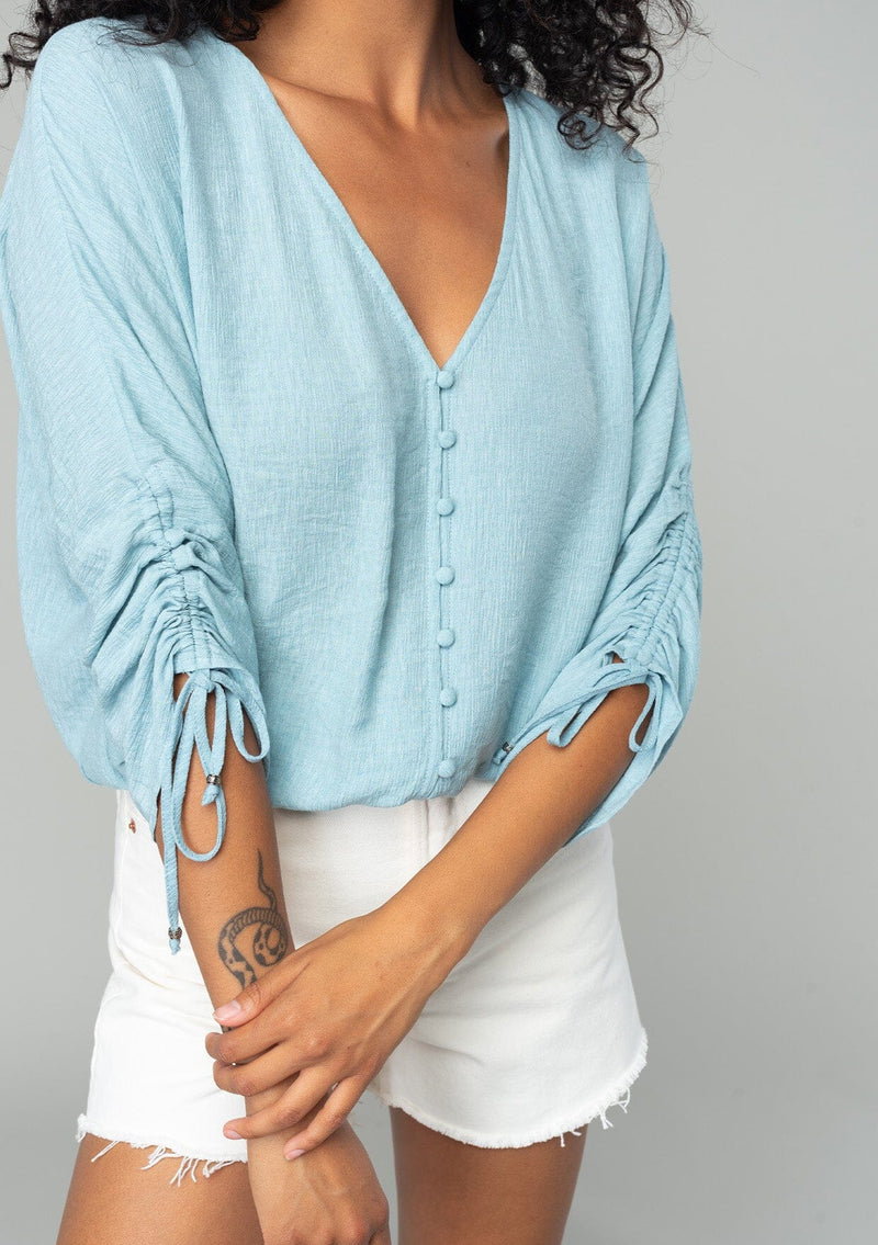 [Color: Dusty Blue] A close up front facing image of a brunette model wearing a classic dusty blue bohemian top with a self covered button front, a v neckline, and three quarter length sleeves with a gathered sleeve detail and adjustable ties. 
