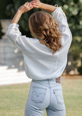 [Color: White] A back facing image of a blonde model wearing a classic white bohemian top with a self covered button front, a v neckline, and three quarter length sleeves with a gathered sleeve detail and adjustable ties.