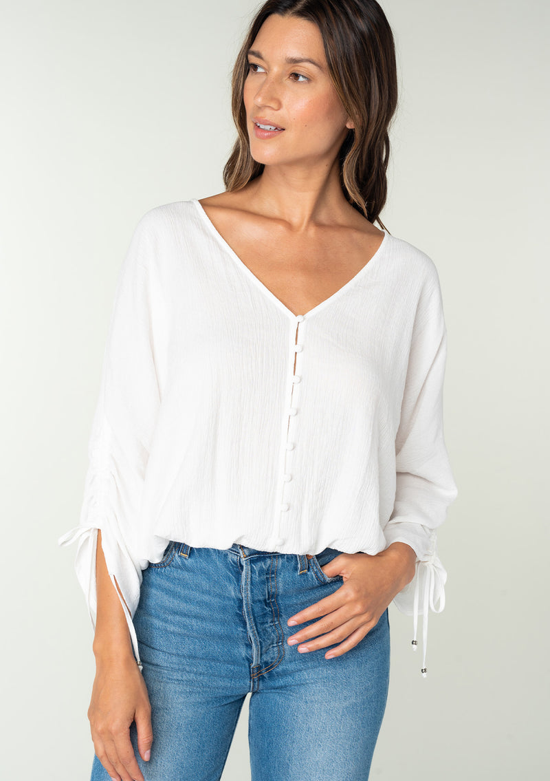 [Color: White] A front facing image of a brunette model wearing a classic white bohemian top with a self covered button front, a v neckline, and three quarter length sleeves with a gathered sleeve detail and adjustable ties. 