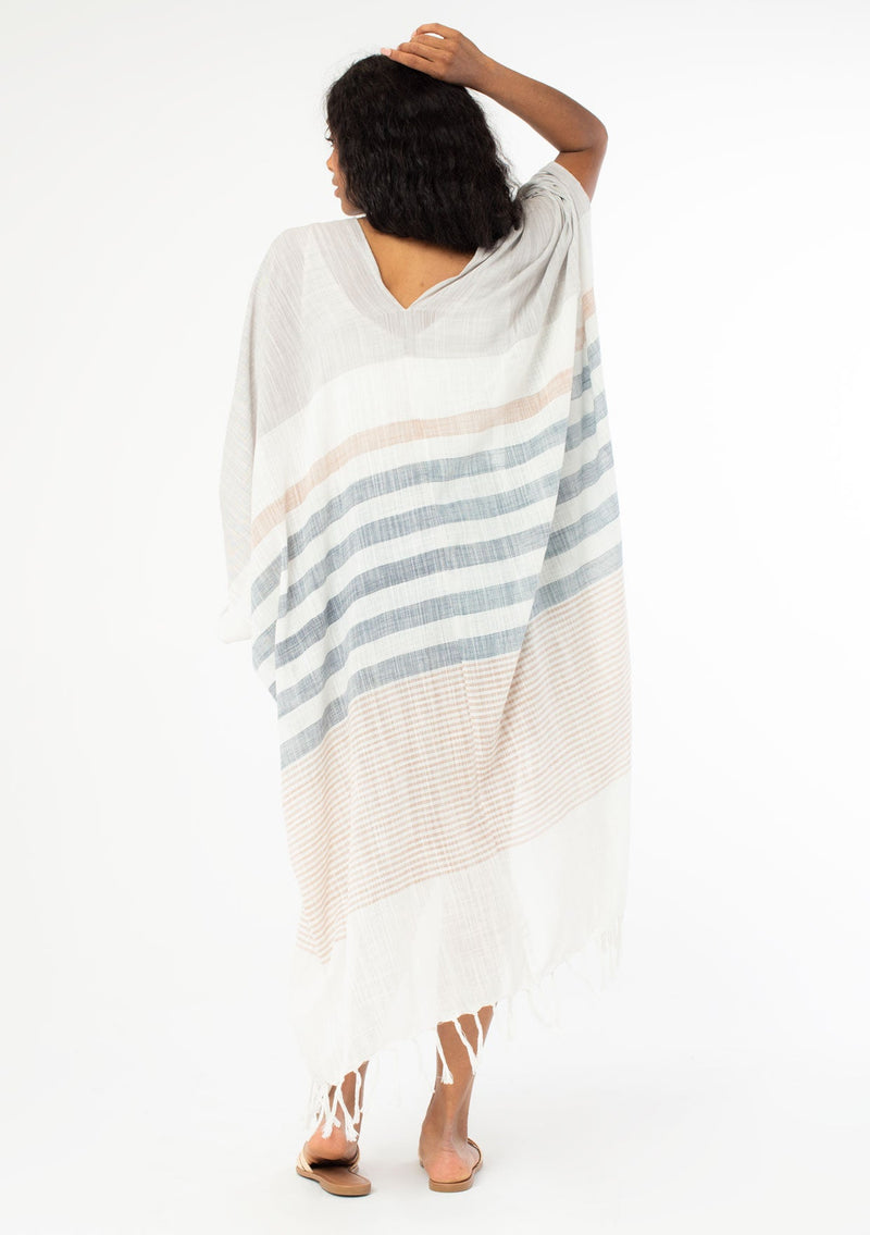 [Color: Neutral Combo] A back facing image of a black model with long wavy hair wearing a cotton bohemian kimono with neutral blue and white white detail and fringe hemline. 