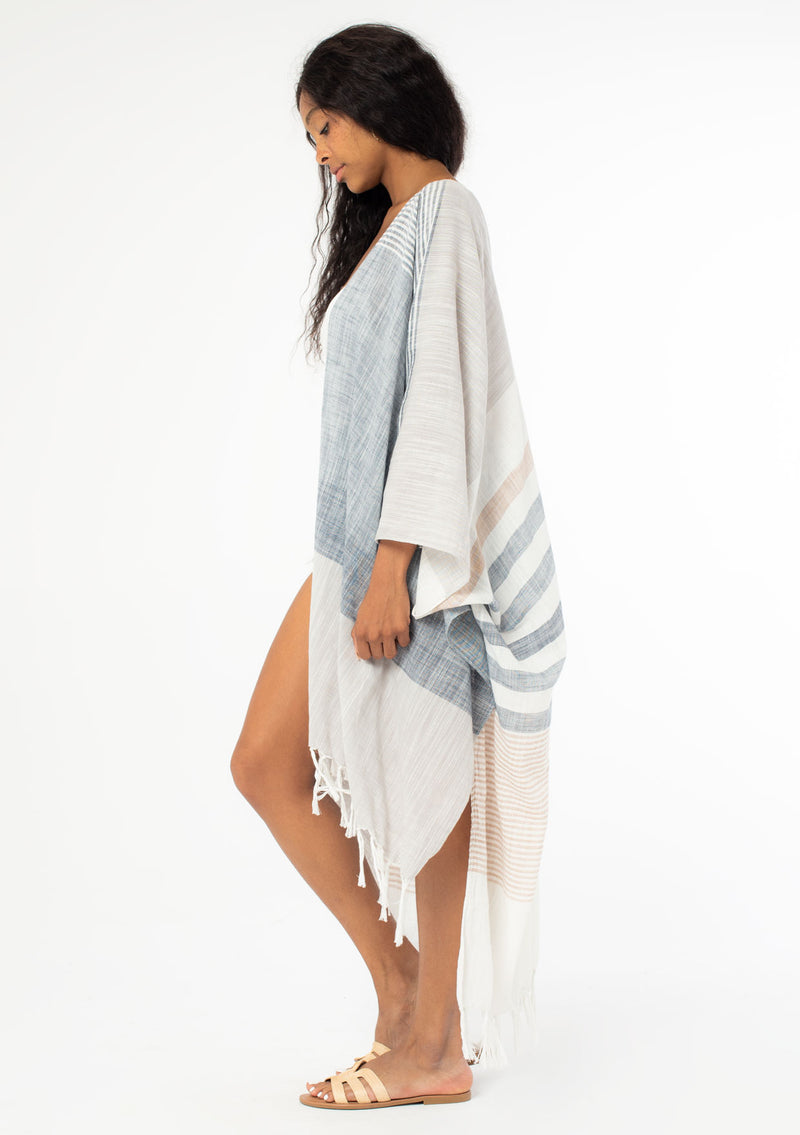[Color: Neutral Combo] A side facing image of a black model with long wavy hair wearing a cotton bohemian kimono with neutral blue and white white detail and fringe hemline. 