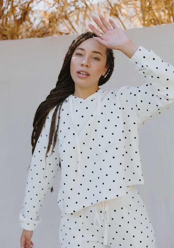 [Color: Ivory/Black] A woman standing outside wearing a cropped polka dot pullover hoodie. Featuring long sleeves with elastic cuffs and a drawstring hoodie. 