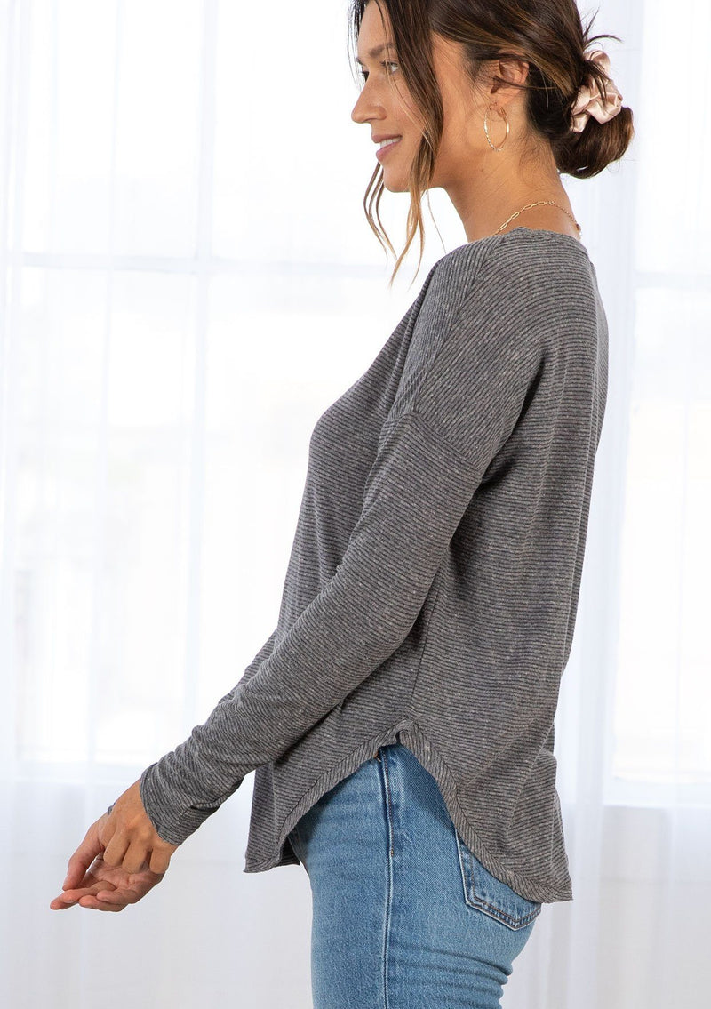 [Color: Charcoal] A model wearing a charcoal cotton jersey stripe long sleeve tee with a wide crew neckline, a dropped shoulder, and a raw hemline. 